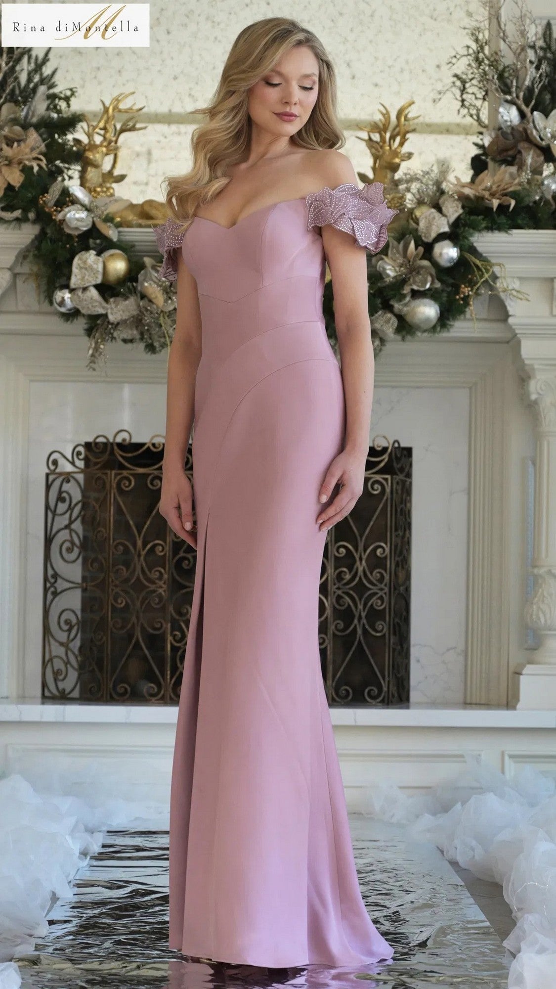 A woman stands indoors in front of a decorated fireplace wearing the Rina Di Montella RD3325 Long Formal Dress in rose, featuring an off-the-shoulder neckline, lace sleeves, and a floor-length sheath silhouette.